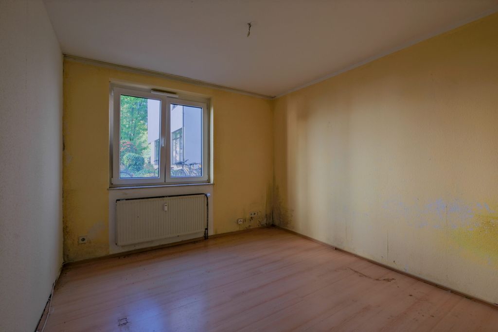 Leerer Raum mit gelben Wänden, einem Fenster mit Blick auf eine Gebäudefassade und einem Heizkörper darunter.