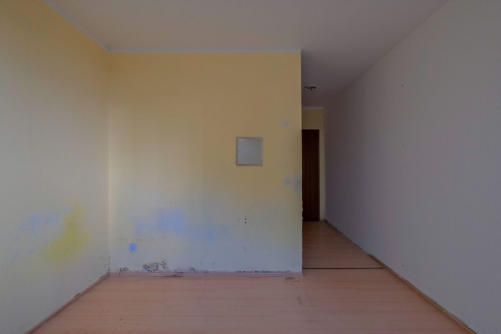Leerer Raum mit gelblich gestrichener Wand und beschädigter Bodenleiste, davor ein heller Holzboden und ein offener Flur mit Tür am Ende.