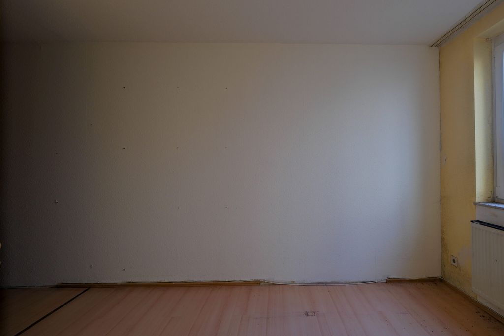 Leerer Raum mit weißen Wänden, Holzfußboden und einem Fenster mit Heizkörper rechts.