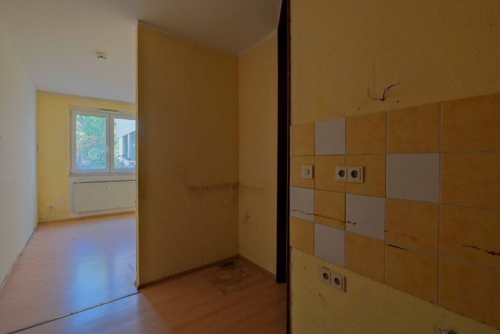 Leerstehender, schmaler Innenraum mit gelb gestrichenen Wänden und einem Fenster mit Blick auf außen, links ist ein Raum mit Laminatboden und Heizkörper sichtbar.