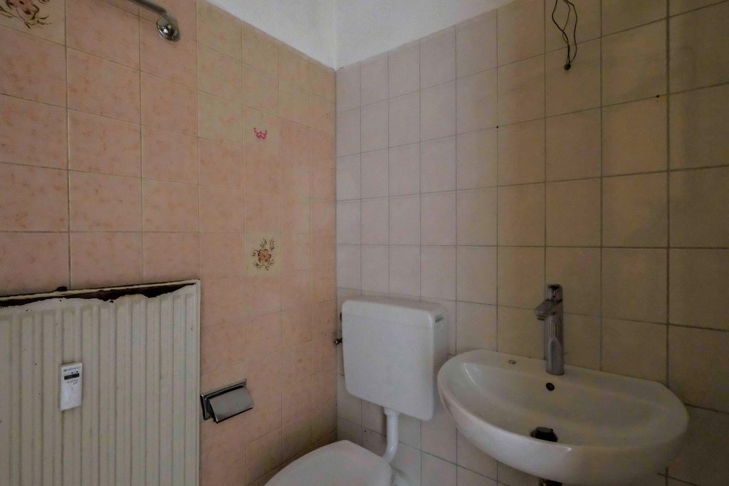 Kleines Badezimmer mit weißen Fliesen, einem weißen Waschtisch mit Wasserhahn, einem WC mit Spülkasten und einem Heizkörper an der Wand.