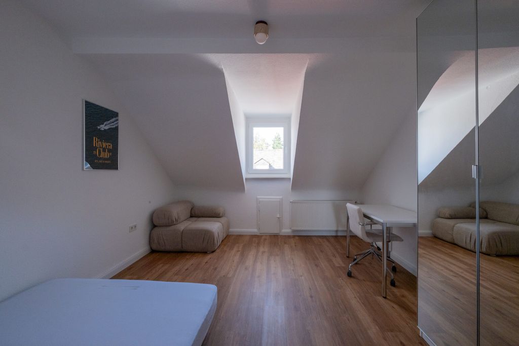 Ein einfach eingerichteter Raum mit einem kleinen Sofa, einem weißen Schreibtisch mit einem Stuhl, einem Bett und einem Fenster in der Dachschräge.