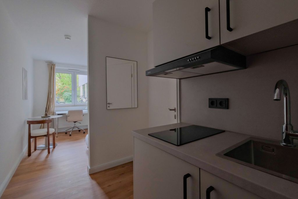 Kleine moderne Küchenzeile mit Herd, Dunstabzugshaube und Spüle, daneben ein heller Raum mit einem runden Tisch, Stuhl und Schreibtisch vor einem Fenster.