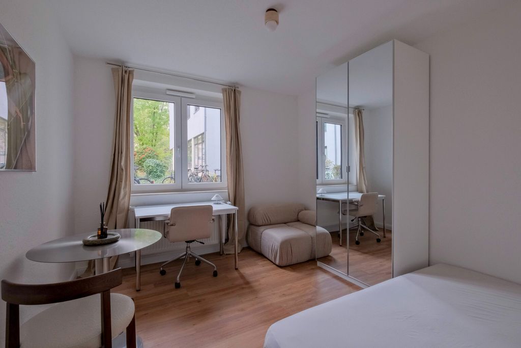 Ein einfach eingerichtetes Schlafzimmer mit einem weißen Bett, einem runden Tisch mit zwei Stühlen, einem Schreibtisch vor einem Fenster mit Blick nach draußen sowie einem beigen Sessel und einem Kleiderschrank mit Spiegeltüren.