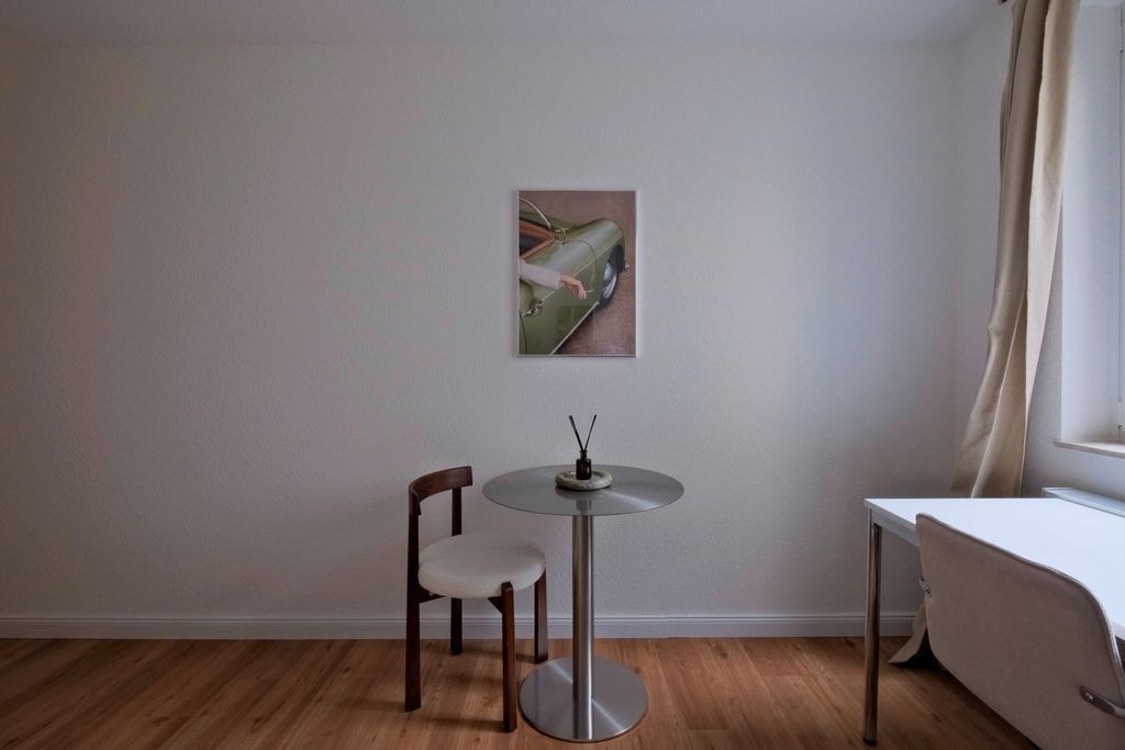 Ein minimalistischer Wohnraum mit einem kleinen runden Metalltisch, einem Stuhl mit weißem Sitzpolster, einem weißen Schreibtisch und einem Bild an der Wand über dem Tisch.