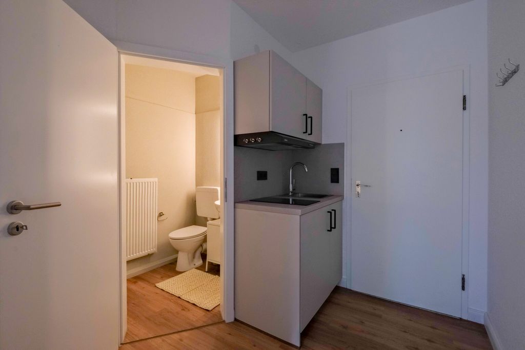 Kleine Küchenzeile mit Spüle und Oberschrank in einem Raum mit weißen Wänden und Holzboden, neben einer offenen Tür zu einem kleinen Bad mit Toilette, Heizkörper und Teppich.