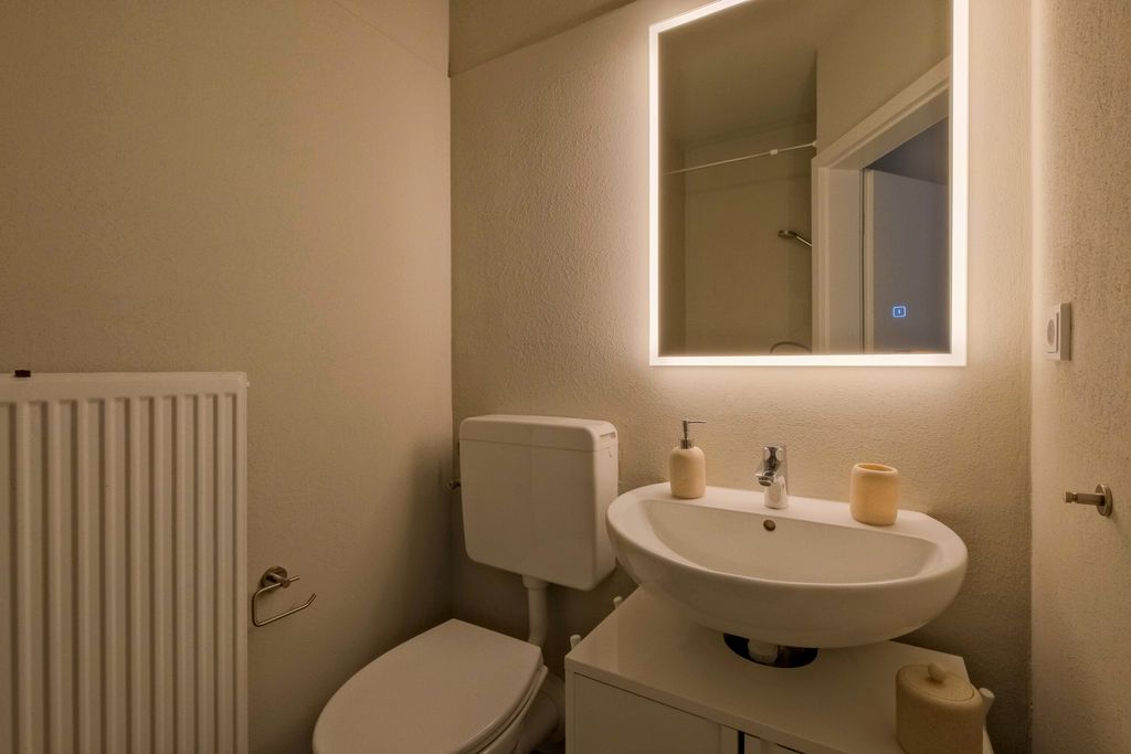 Kleines Badezimmer mit einer Toilette, einem Waschbecken mit Seifenspender und Behälter sowie einem beleuchteten Spiegel an der Wand.