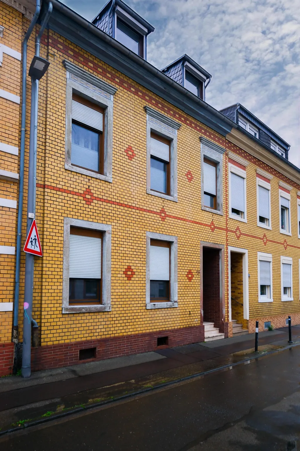 Wohngebäude mit gelbem und rotbraunem Backsteinmuster, mehreren Fenstern mit weißen Rollläden und Dachgauben unter bewölktem Himmel.