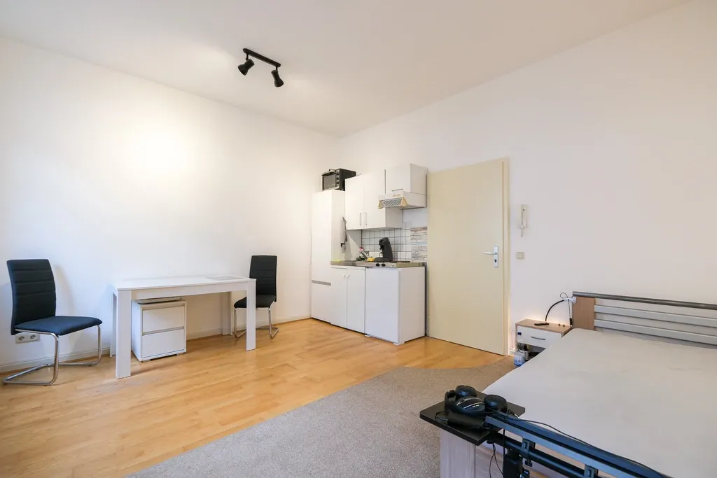 Moderner, heller Wohnraum mit Küchenzeile, zwei schwarzen Stühlen, Schreibtisch und Bett mit grauer Bettwäsche.