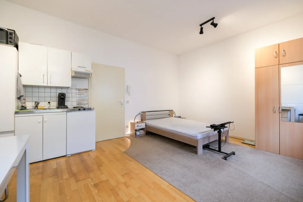 Ein einfach eingerichtetes Zimmer mit einem Einzelbett auf einem grauen Teppich, einem kleinen Nachttisch mit Lampe, einer Küchenzeile mit weißer Möblierung und einem großen Holzkleiderschrank mit eingebautem Spiegel.