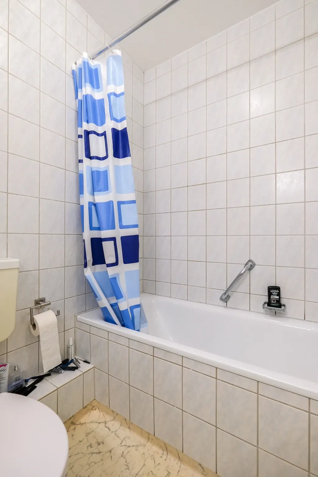 Ein Badezimmer mit weiß gefliester Badewanne, einer blau karierten Duschvorhang, einer Toilettenpapierrolle und einer Zahnbürste auf der Ablage neben der Toilette.