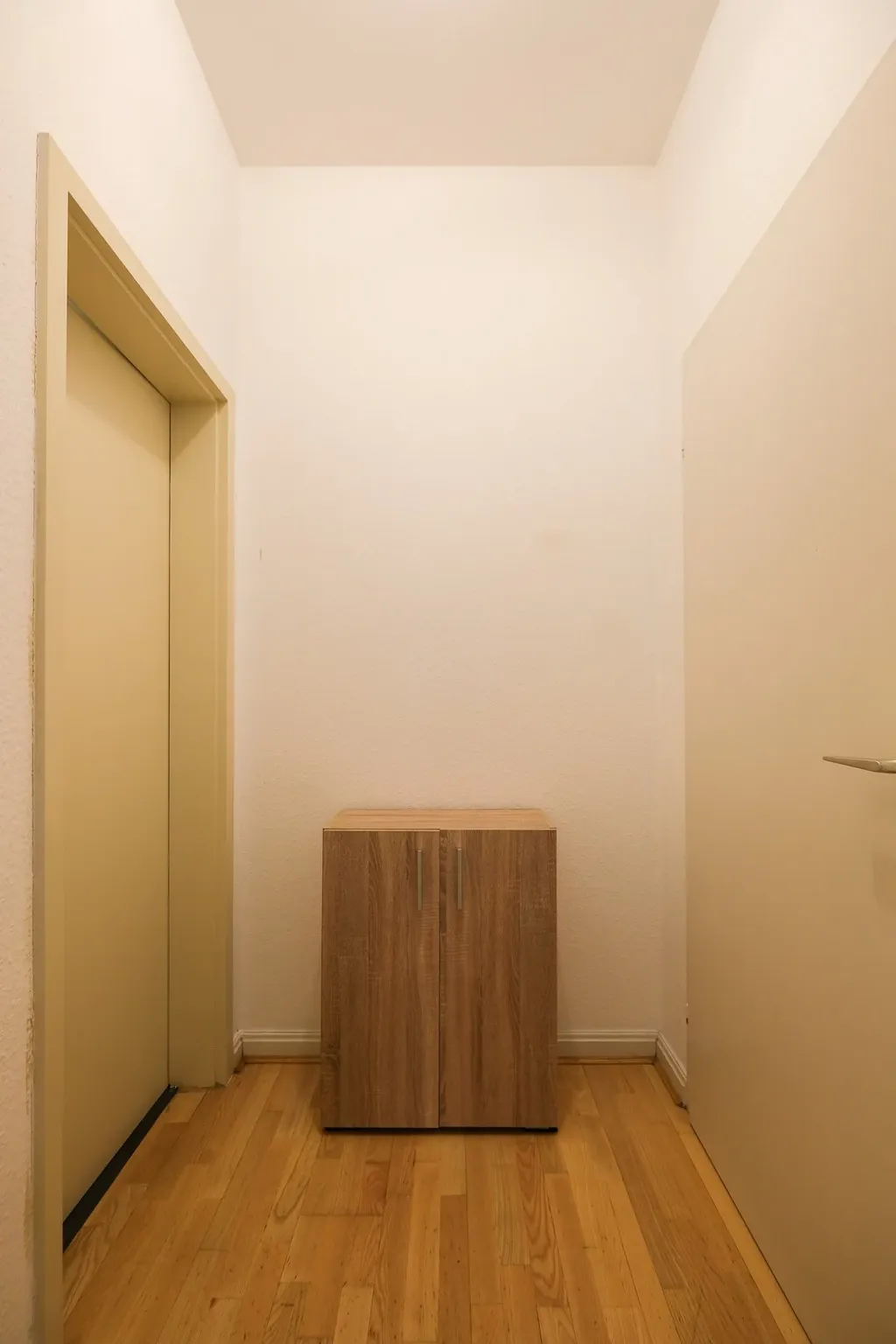 Schmaler Hausflur mit Holzboden, links eine gelbe Tür, rechts eine weiße Tür mit silbernem Türgriff und mittig ein kleines zweiflügeliges Holzschränkchen.