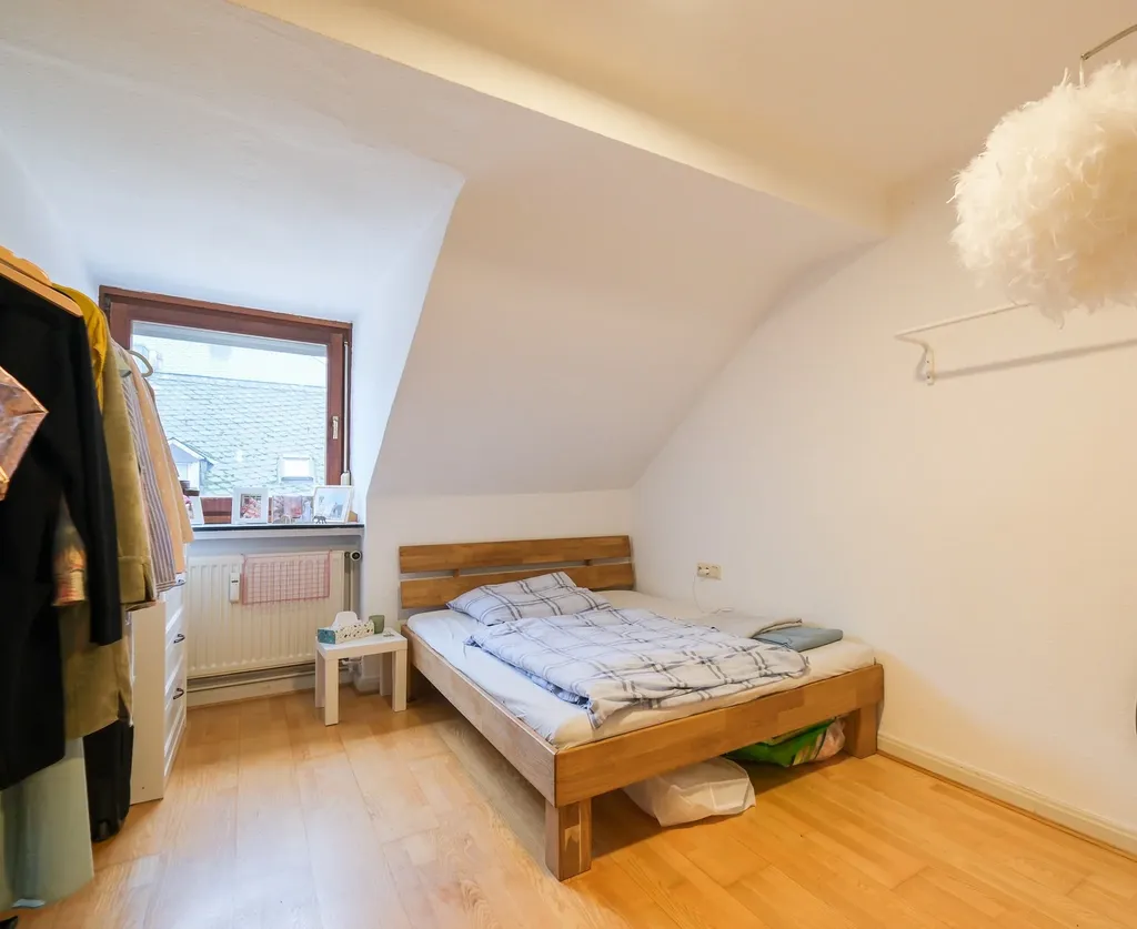 Schlafzimmer mit Holzlattenbett, hellen Holzboden, Fenster mit Blick auf Dachziegel und Kleiderschrank mit Kleidung.