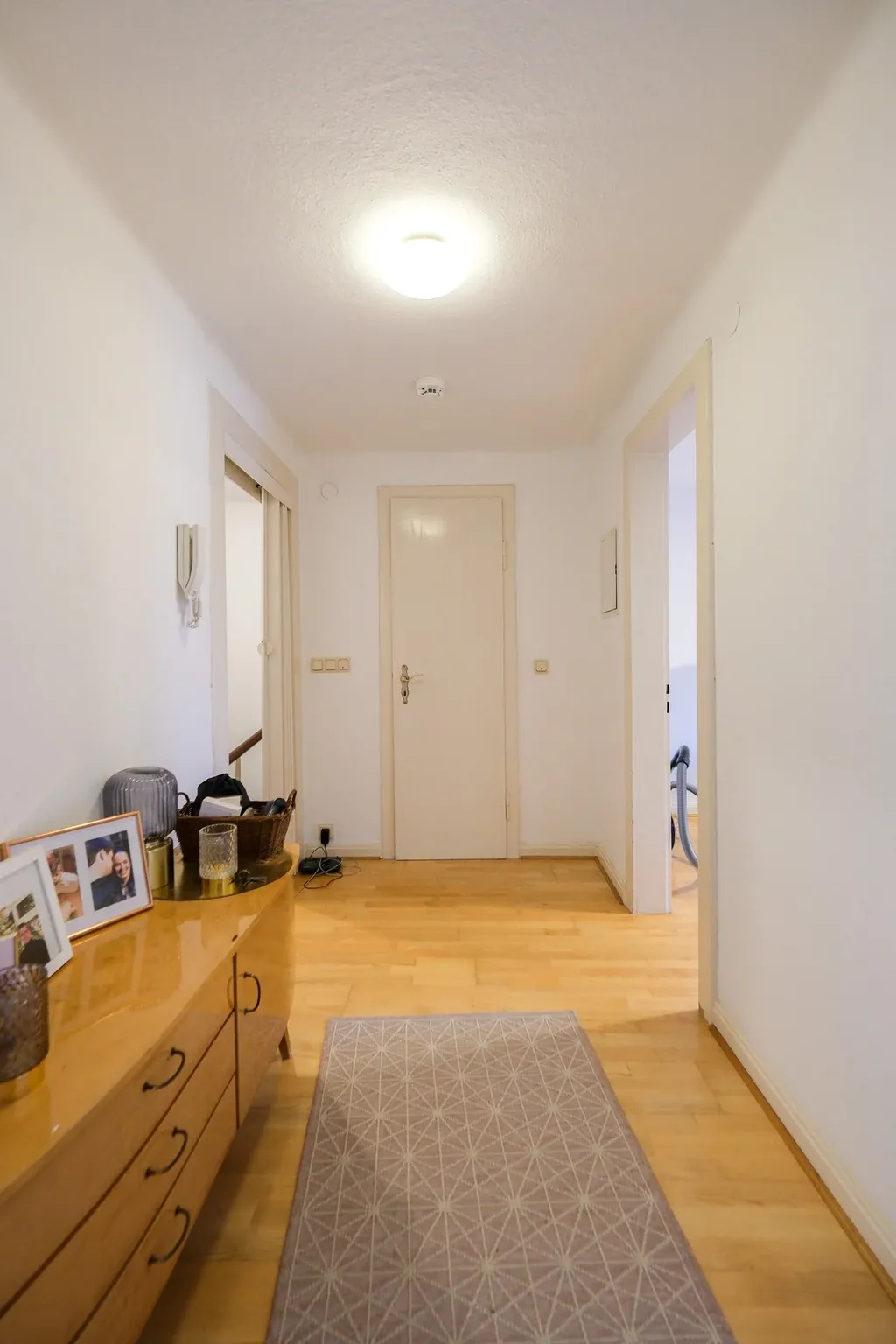 Ein heller Flur mit hellen Holzfußböden, einem beigen Teppich in der Mitte, weiß gestrichenen Wänden und einer geschlossenen Tür am Ende. Links steht eine Kommode mit Familienfotos und dekorativen Gegenständen.