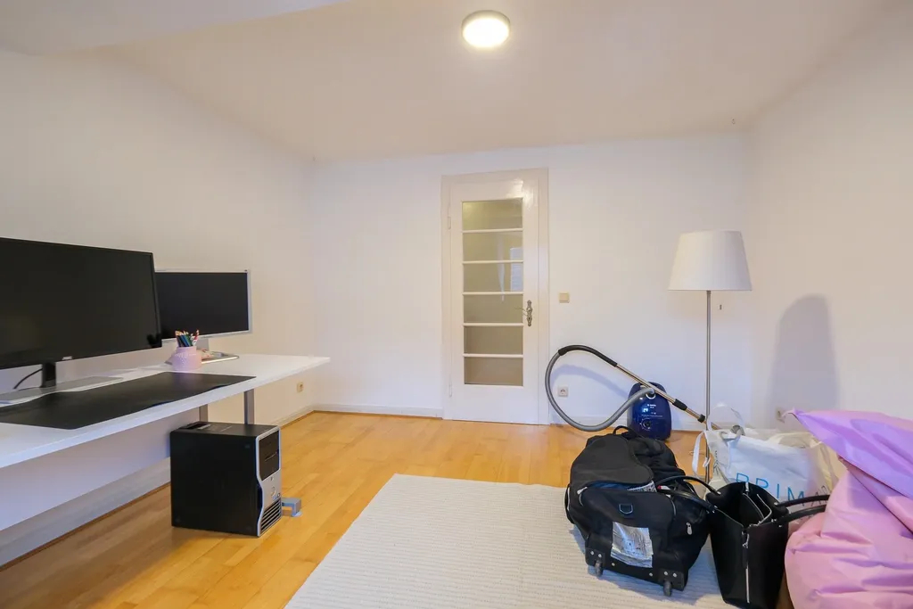 Ein Wohnraum mit weißem Schreibtisch und zwei Monitoren auf der linken Seite, einem weißen Teppich auf Holzboden und einer weißen Tür im Hintergrund sowie einem Staubsauger, einer Stehlampe und Koffern auf der rechten Seite.