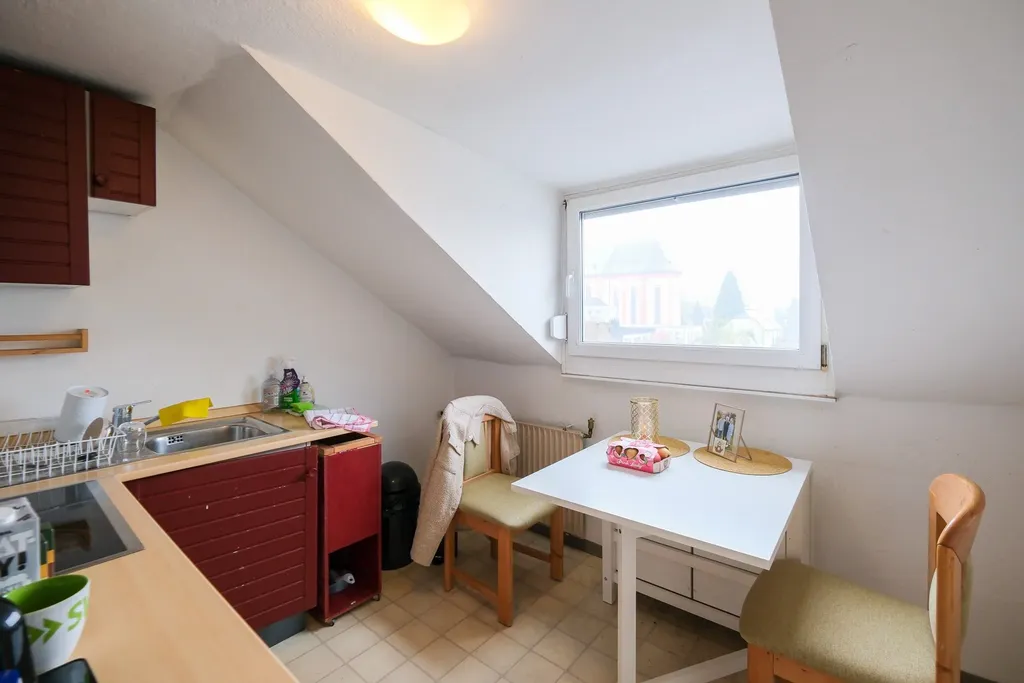 Kleine Küche mit rotem Schrank, Spüle, zwei Stühlen und einem weißen Tisch unter einem Dachfenster.