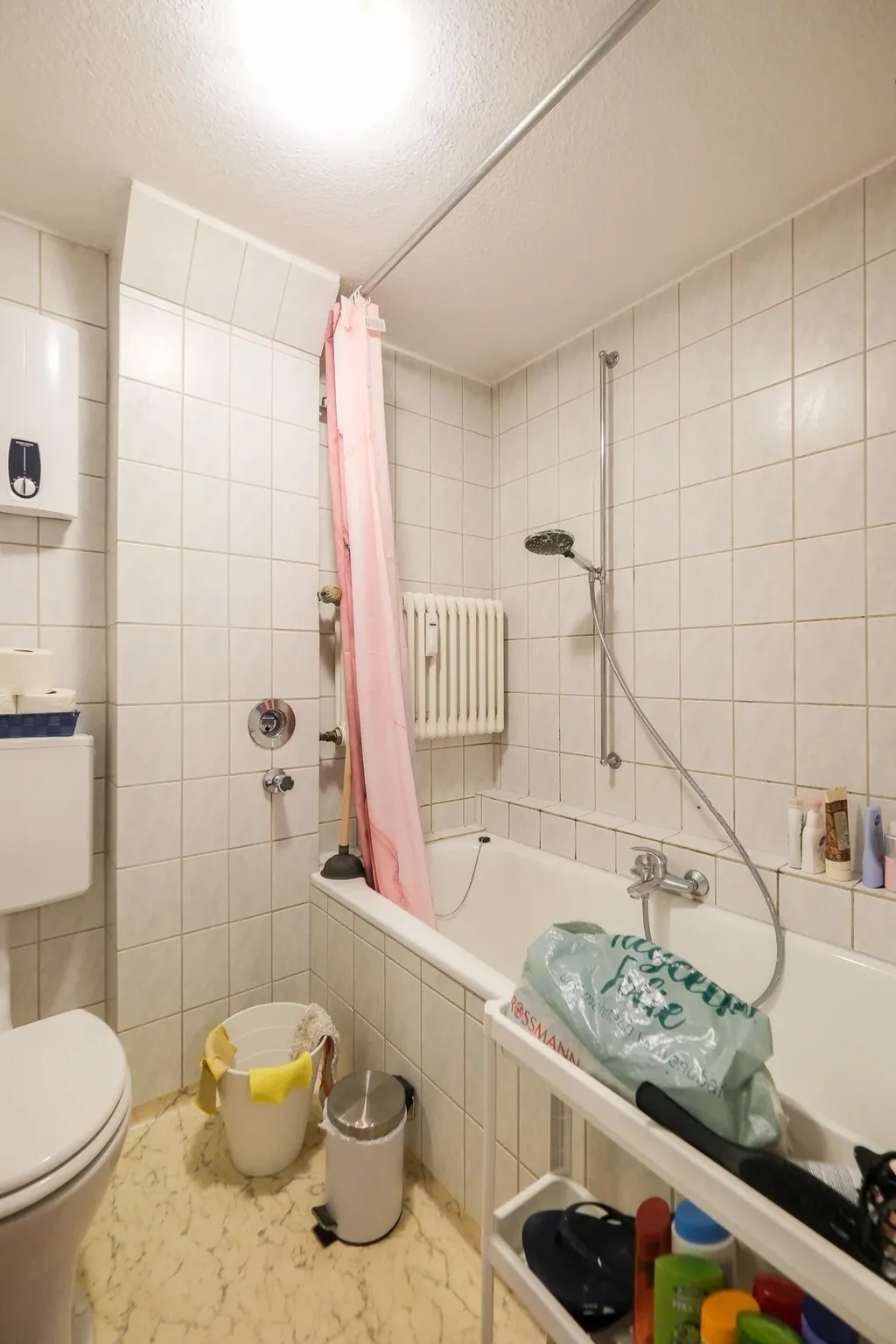 Ein weiß gekacheltes Badezimmer mit Badewanne, rosa Duschvorhang, einem Eimer mit gelben Handschuhen, einem Mülleimer und einer Toilette.
