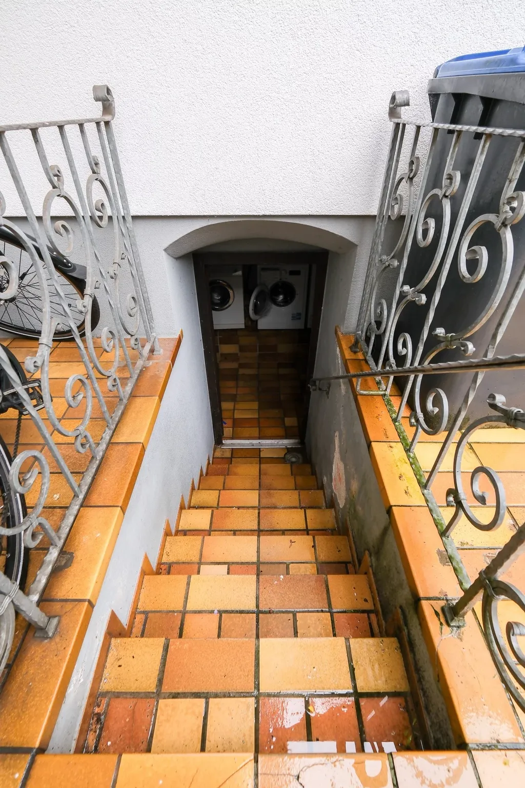 Eine Treppe mit orangefarbenen Fliesen führt zu einem Kellerraum mit zwei Waschmaschinen, umgeben von dekorativem Geländer und einer Mülltonne.