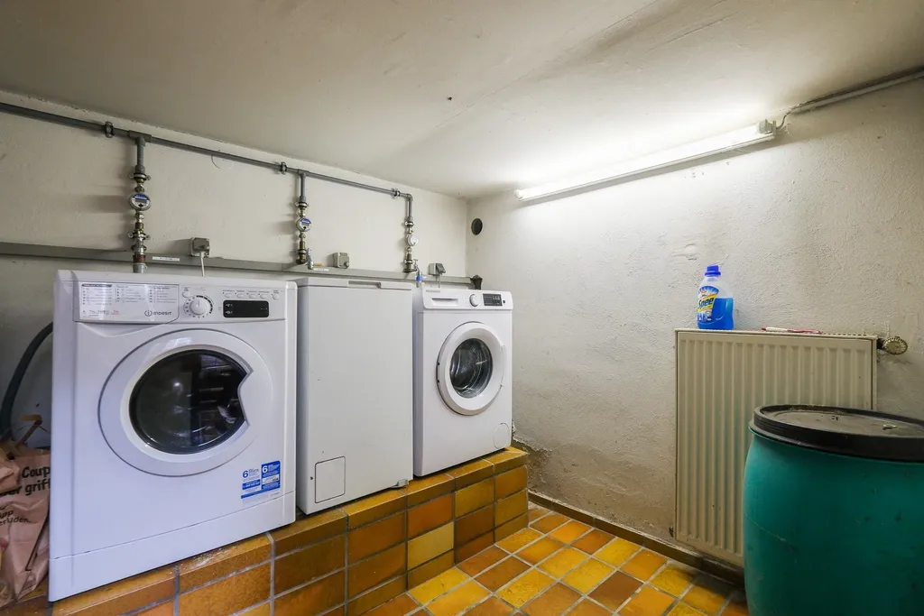Ein Kellerraum mit zwei weißen Waschmaschinen und einem Kühlschrank auf einer erhöhten, gekachelten Plattform, daneben ein Heizkörper mit einer Reinigungsflasche darauf und ein großer blauer Behälter.