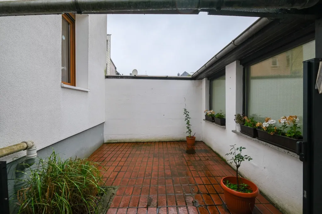 Kleiner, leerer Innenhof mit rotem Pflaster, weißen Wänden, einigen Pflanzen in Töpfen und Blumen in Fensterkästen an einem Gebäudefenster.