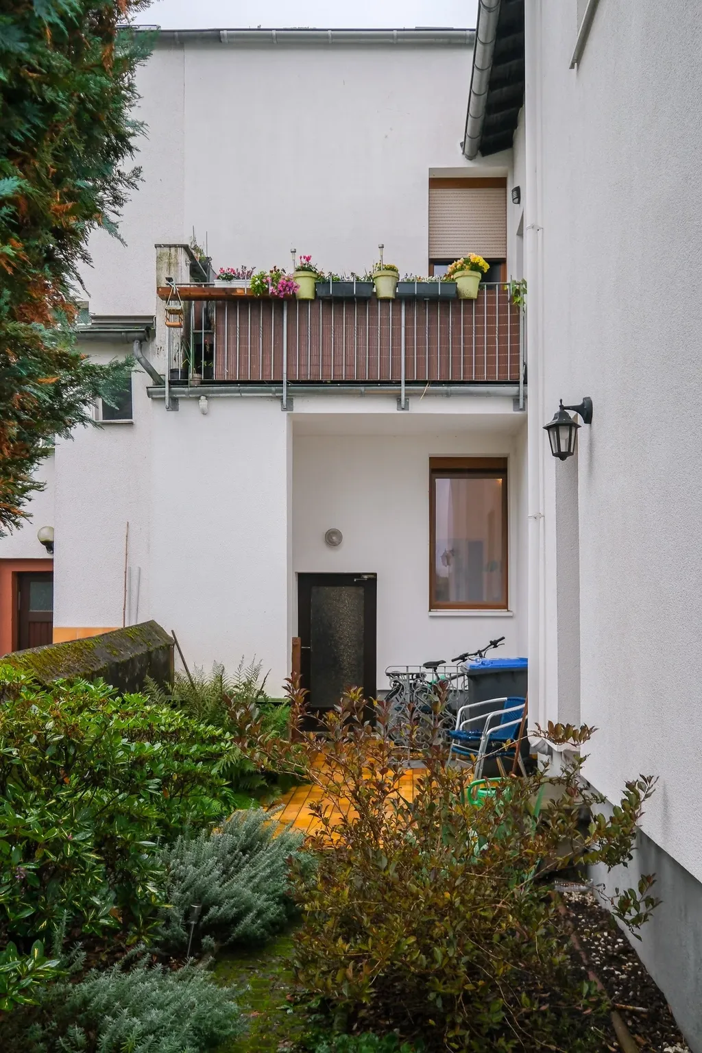 Der Eingangsbereich eines weißen Mehrfamilienhauses mit Balkon, auf dem Blumenkästen stehen, umgeben von dichtem Grün.