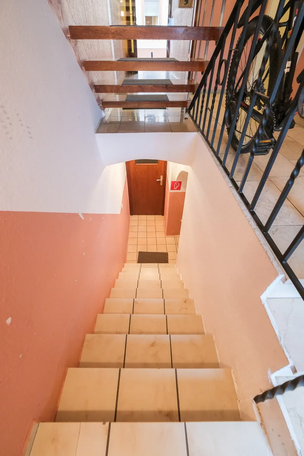 Blick von oben auf eine nach unten führende Treppe mit weißen Fliesen, umgeben von weißen und rosa Wänden, mit einer Holztür am unteren Ende und einem Fahrrad neben dem Geländer im oberen Bereich.