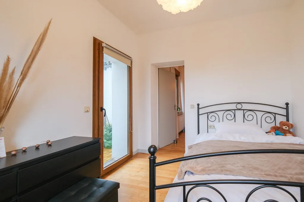 Helles Schlafzimmer mit einem schwarzen Metallbett, einem Teddy auf dem Bett und einer Kommode neben einer Tür mit Glasfenster zum Außenbereich.