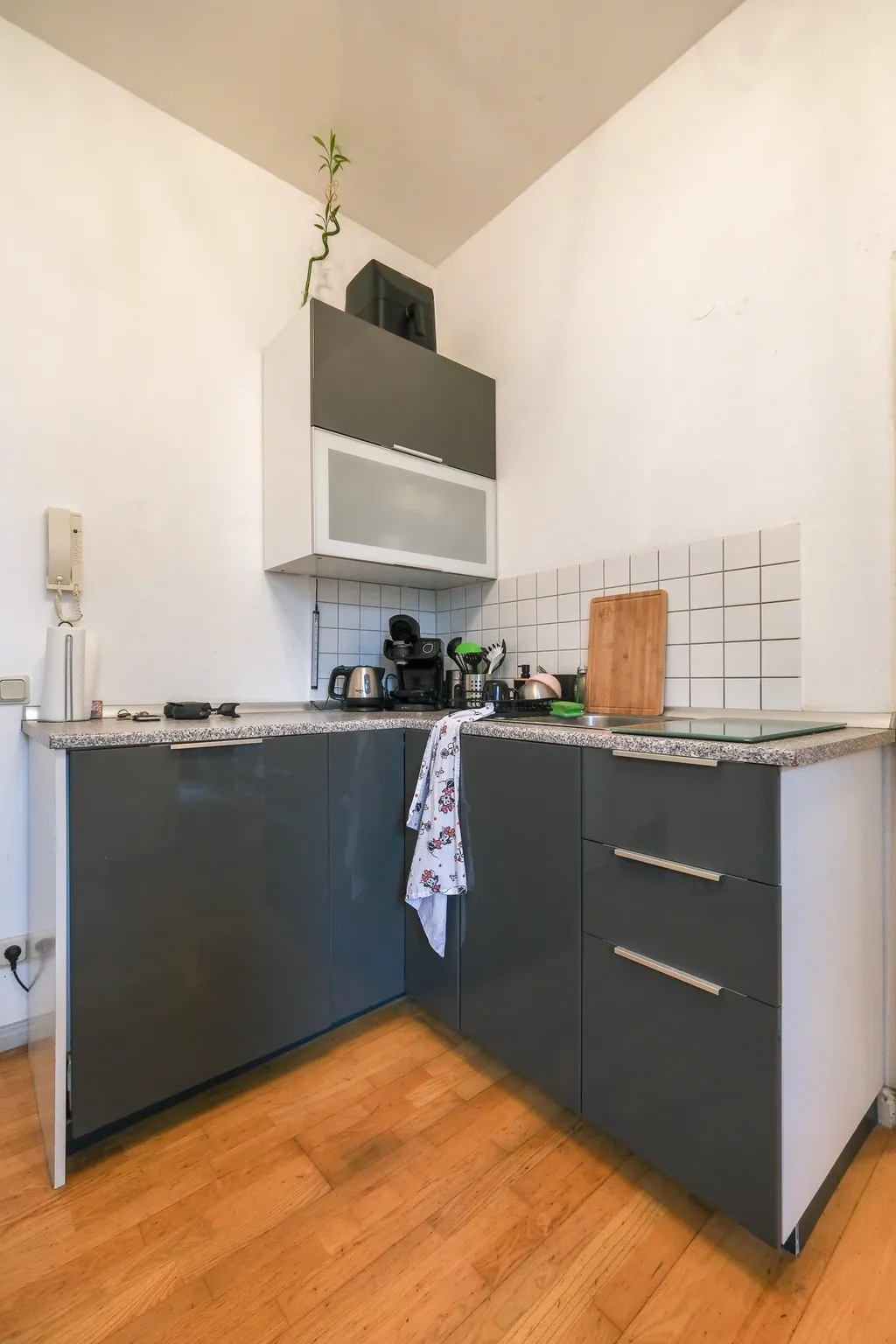 Kleine moderne Küche mit grauen Schränken, Holzfußboden und weißen Wänden, ausgestattet mit Kaffeemaschine, Toaster, Schneidebrett und Geschirrtuch.