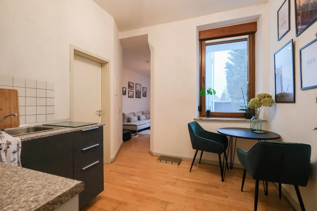 Moderner Küchenbereich mit grauen Schränken, grauer Arbeitsplatte und Spülbecken, daneben ein heller Flur mit Blick auf ein Wohnzimmer mit Sofa und einige Bildrahmen an der Wand.