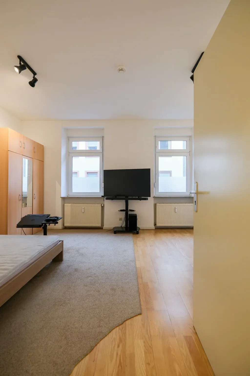 Ein schlicht eingerichtetes Schlafzimmer mit einem Bett ohne Bettwäsche, einem großen Teppich, einem Kleiderschrank mit Spiegeltüren und einem Fernseher vor zwei Fenstern.