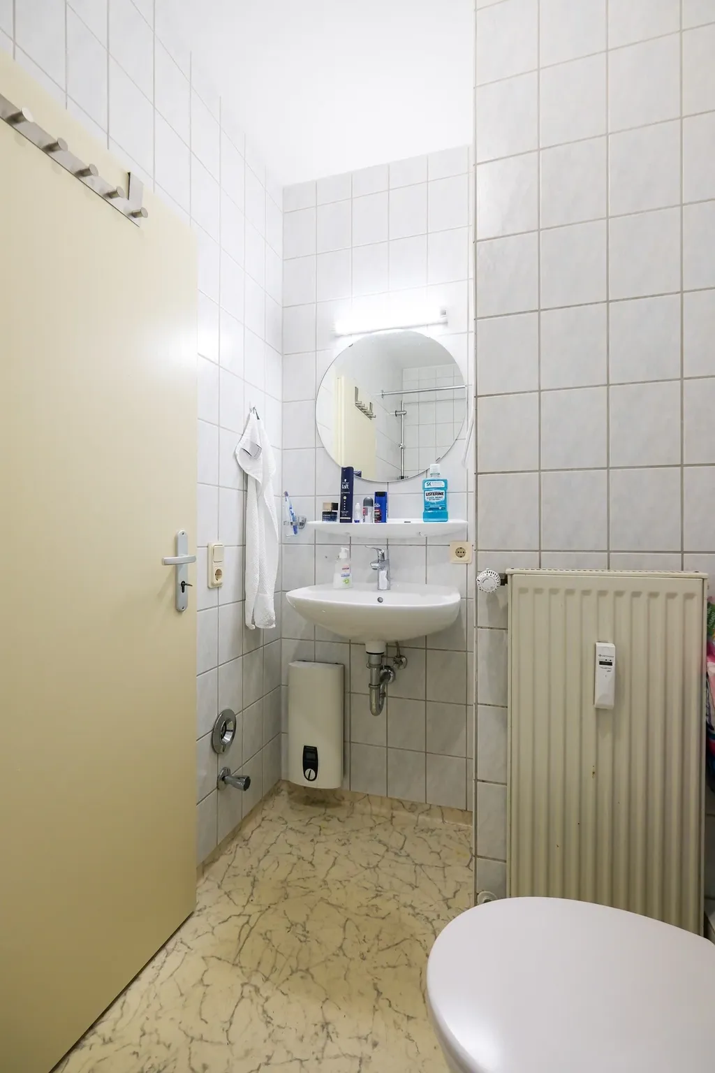 Kleines Badezimmer mit weißem Waschbecken, rundem Spiegel, weißen Kacheln und hellbraunem Marmorboden. An der Wand hängen ein Handtuch und verschiedene Flaschen mit Hygieneprodukten.