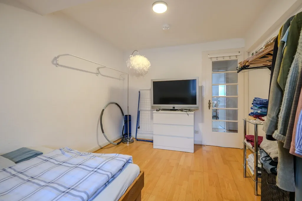 Schlafzimmer mit Holzboden, einem Bett mit blau kariertem Bettbezug, einem Fernseher auf einer weißen Kommode und Kleidung auf einem offenen Kleiderständer.