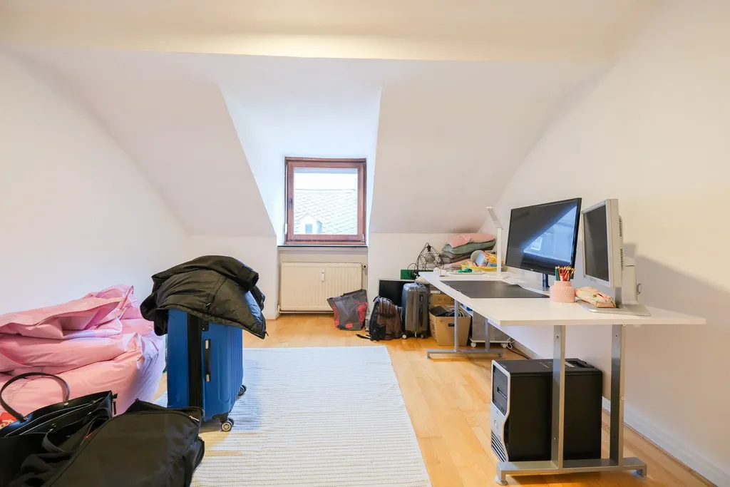 Heller Dachgeschossraum mit Schrägen, Fenster und Holzboden, mit Arbeitsbereich bestehend aus zwei Monitoren und PC-Tower sowie Koffern und Kartons im Hintergrund.