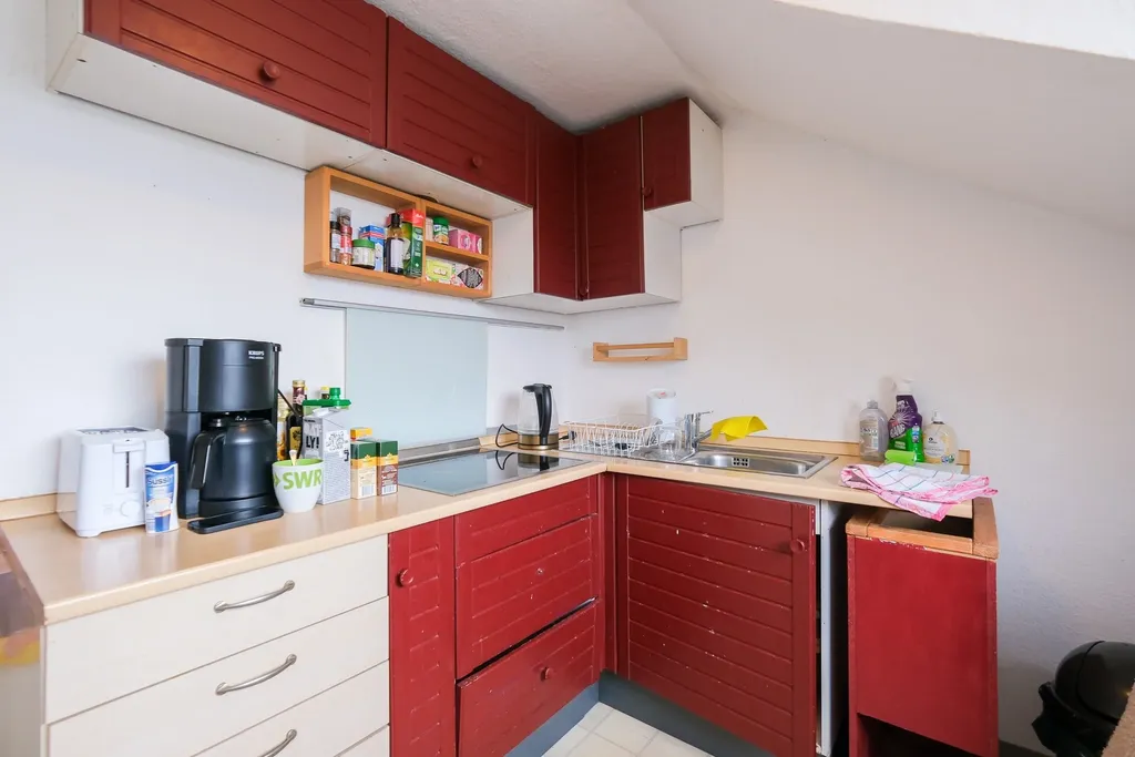 Kleine, moderne Küche mit roten Schränken, Kaffeemaschine, Spüle und Kochfeld unter einer schrägen Decke.
