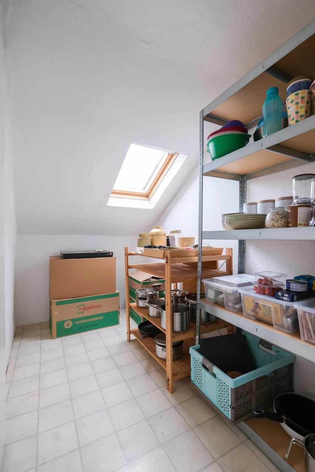 Ein Vorratsraum mit schrägem Dachfenster, Regalen aus Metall und Holz, die mit Töpfen, Plastikbehältern und Kartons bestückt sind.