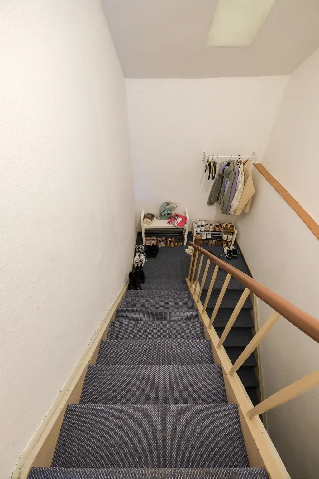 Treppe mit blaugrauem Teppich, am unteren Ende Schuhregale und eine Wand mit aufgehängten Jacken in einem Innenbereich.