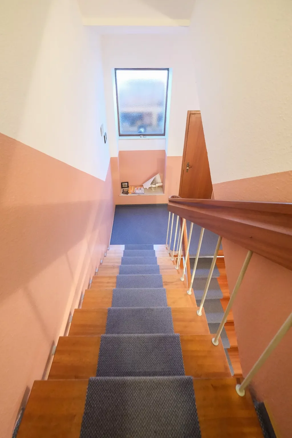 Treppe mit dunkelblauem Teppichläufer, beigen Wänden und einem Fenster mit Milchglas am Treppenabsatz neben einer Holztür.