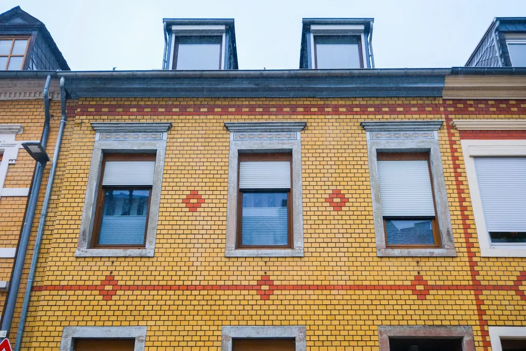 Fassade eines gelben Backsteinhauses mit drei Fenstern und Dachgauben unter einem blauen Himmel.