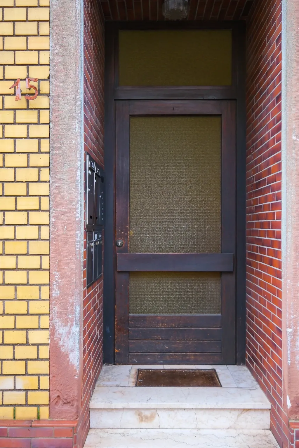 Eingangstür eines Gebäudes mit gelben und roten Ziegelwänden, Hausnummer 15 links an der Wand, mehrere Briefkästen neben der Tür, zwei Marmorstufen führen zur dunklen Tür.