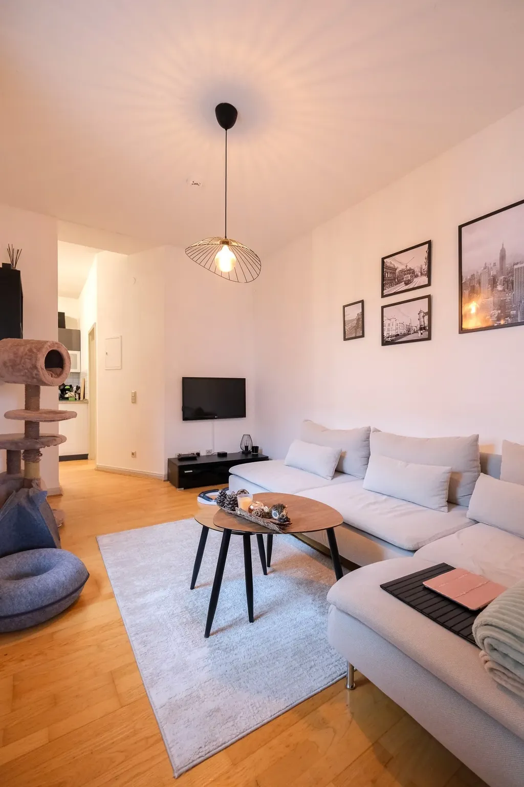 Helles Wohnzimmer mit einer großen grauen Eckcouch, zwei ineinandergestellten Holztischen auf einem hellen Teppich, Wandbildern und einem Katzenkletterbaum neben der Couch.