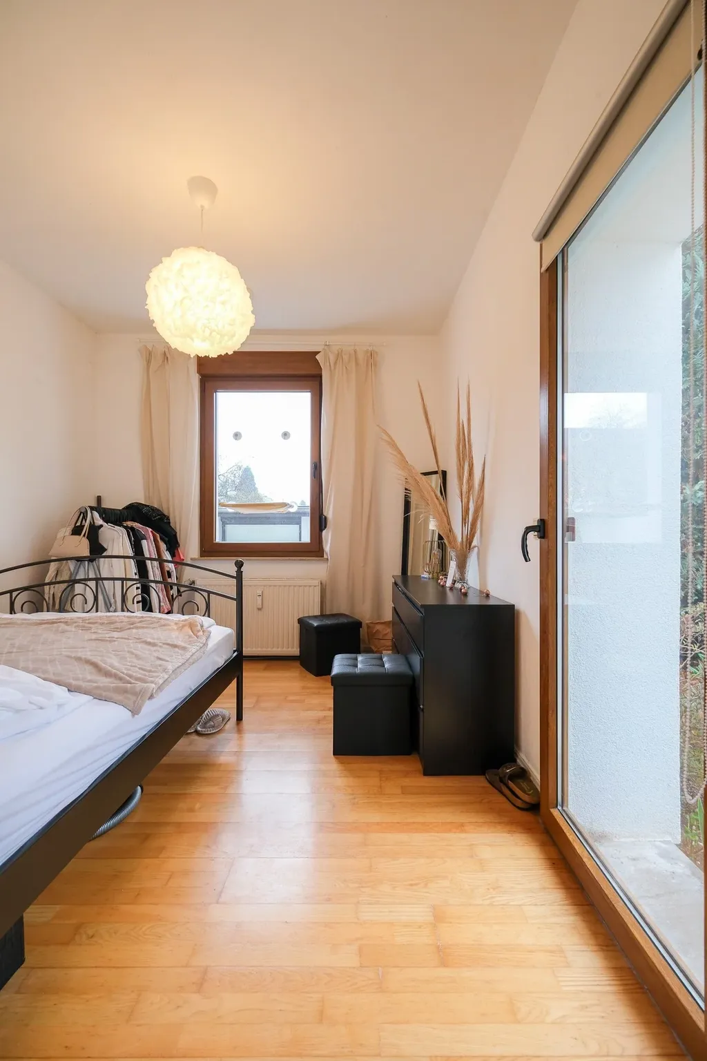 Hell eingerichtetes Schlafzimmer mit einem Metallbett, einem Fenster mit beigen Vorhängen, einem schwarzen Schrank mit dekorativen Pflanzen und einem großen Fenster mit Sicht auf einen Balkon.