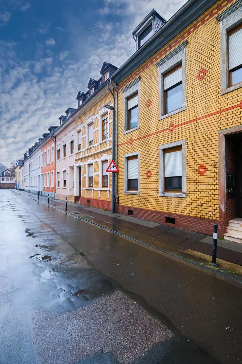 Reihe von farbigen Wohnhäusern mit gelben Ziegelwänden und weißen Fensterläden an einer nassen Straße unter einem bewölkten Himmel.