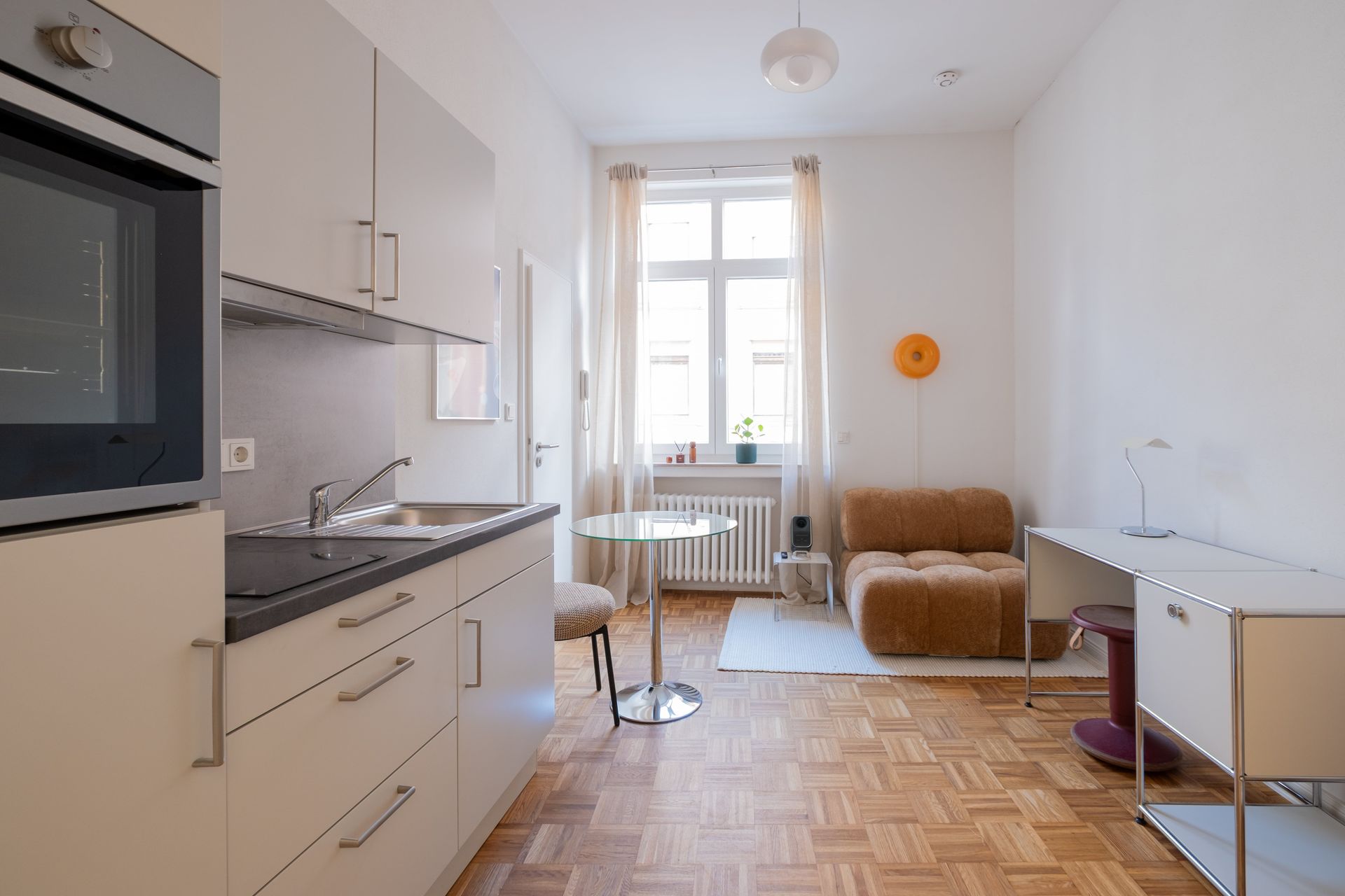 Moderne helle Einzimmerwohnung mit Küchenzeile, Glas-Essbereich, braunem Sessel und schlichtem Schreibtisch vor einem großen Fenster.