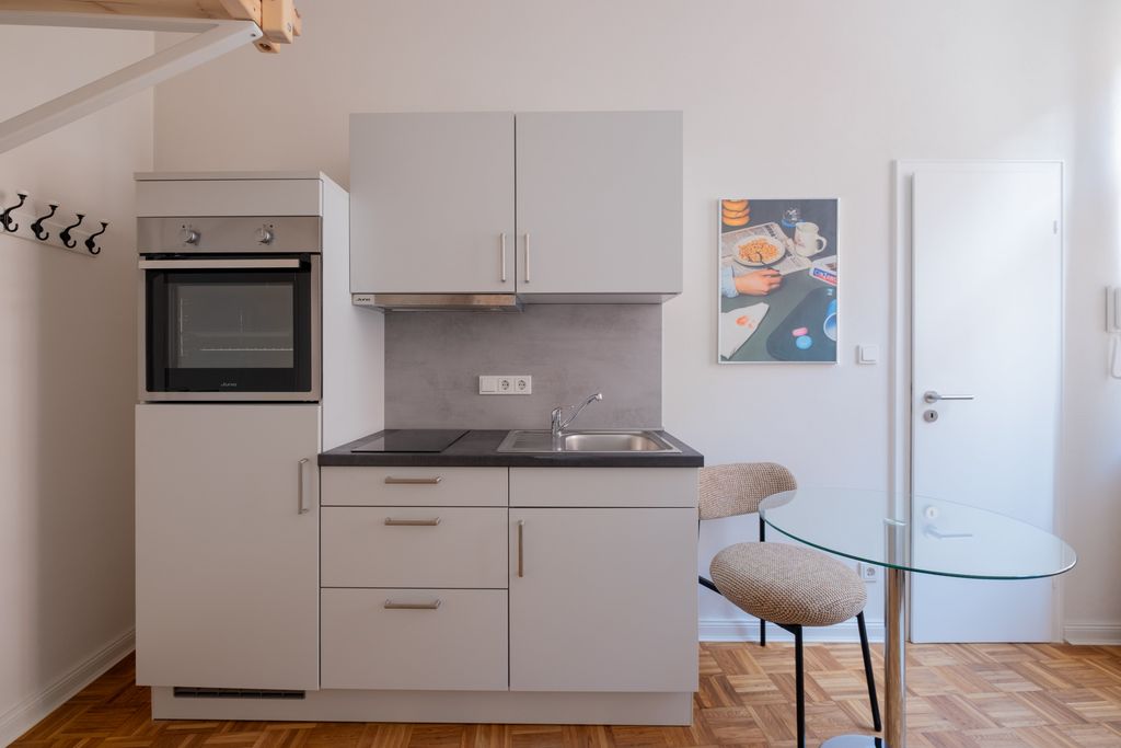 Kleine moderne Küchenzeile mit grauen Schränken, eingebautem Ofen, Herd, Spüle und einem Glastisch mit zwei gepolsterten Stühlen in einem hellen Raum.