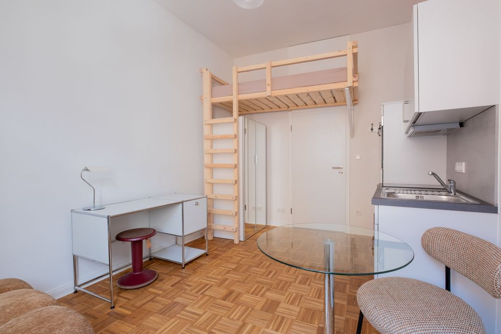 Kleines Apartment mit Holzboden, weißem Schreibtisch mit rotem Hocker, hochgelegtem Bett aus Holz und Küchenzeile mit Spüle und Stuhl.