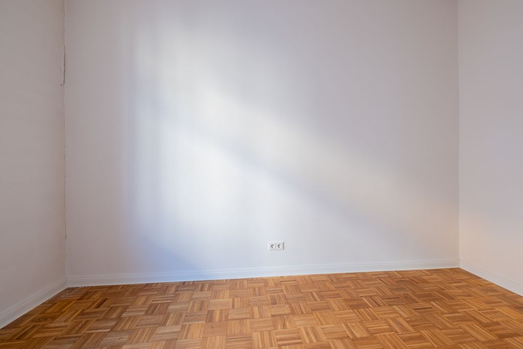 Leerer Raum mit hellem Parkettboden und weißen Wänden, Sonnenlichtschein fällt auf eine Wand mit Steckdosen.