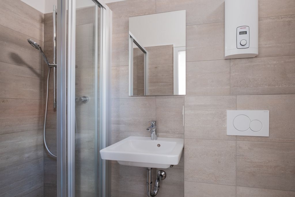 Kleines Badezimmer mit Dusche, Waschbecken, Spiegel, Wandheizung und Toilette mit Wandspülkasten in hell gefliesten Wänden.