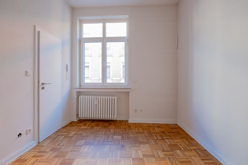 Leerer Raum mit weiß gestrichenen Wänden, einem Fenster mit Blick auf ein Nachbargebäude, einer Heizung unter dem Fenster und einem weißen Türblatt auf der linken Seite.