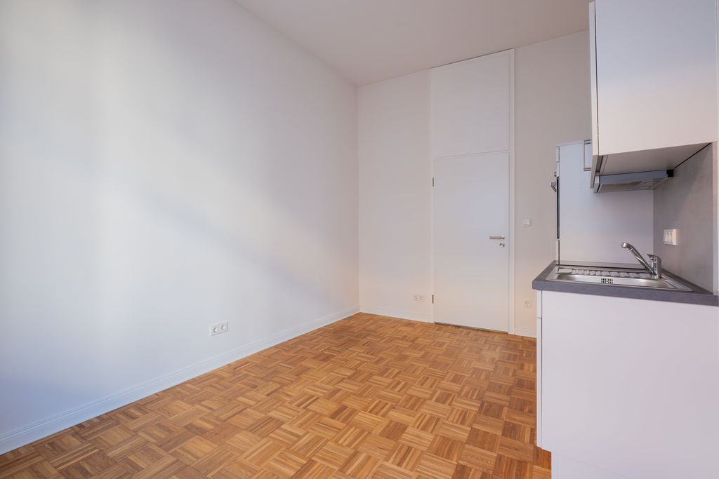 Leerer Raum mit weiß gestrichenen Wänden, Holzparkettboden und einer kleinen Küchenzeile mit Spüle auf der rechten Seite.