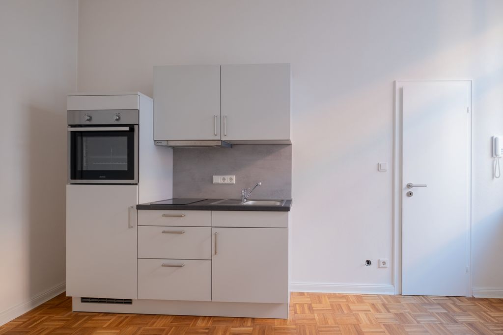 Kleine Küchenzeile in einem leeren, schlicht gehaltenen Raum mit weißer Wand, Holzboden und geschlossener Tür rechts.